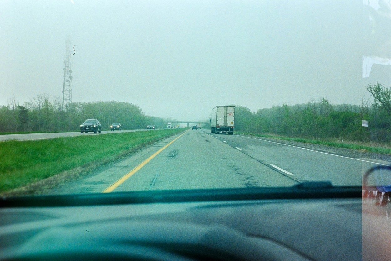 A view from inside a vehicle driving on a multi-lane highway on a foggy, overcast day. A large white semi-truck travels ahead in the right lane, while several cars can be seen approaching in the oncoming lanes to the left. The road is bordered by green grass and trees, with a tall communication tower visible on the left side. Yellow and white lane markings are present, and the road surface appears slightly wet.